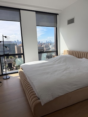 Quarto com cama grande, vista da cidade através de janelas do chão ao teto e abajur moderno.