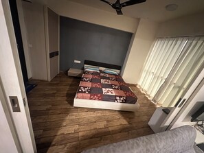 Un dormitori amb un llit, un ventilador de sostre, una finestra amb cortines i un terra de fusta.