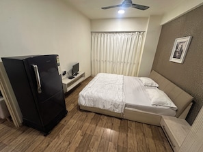 A compact hotel room with a bed, a small desk with a computer, a refrigerator, and a ceiling fan.