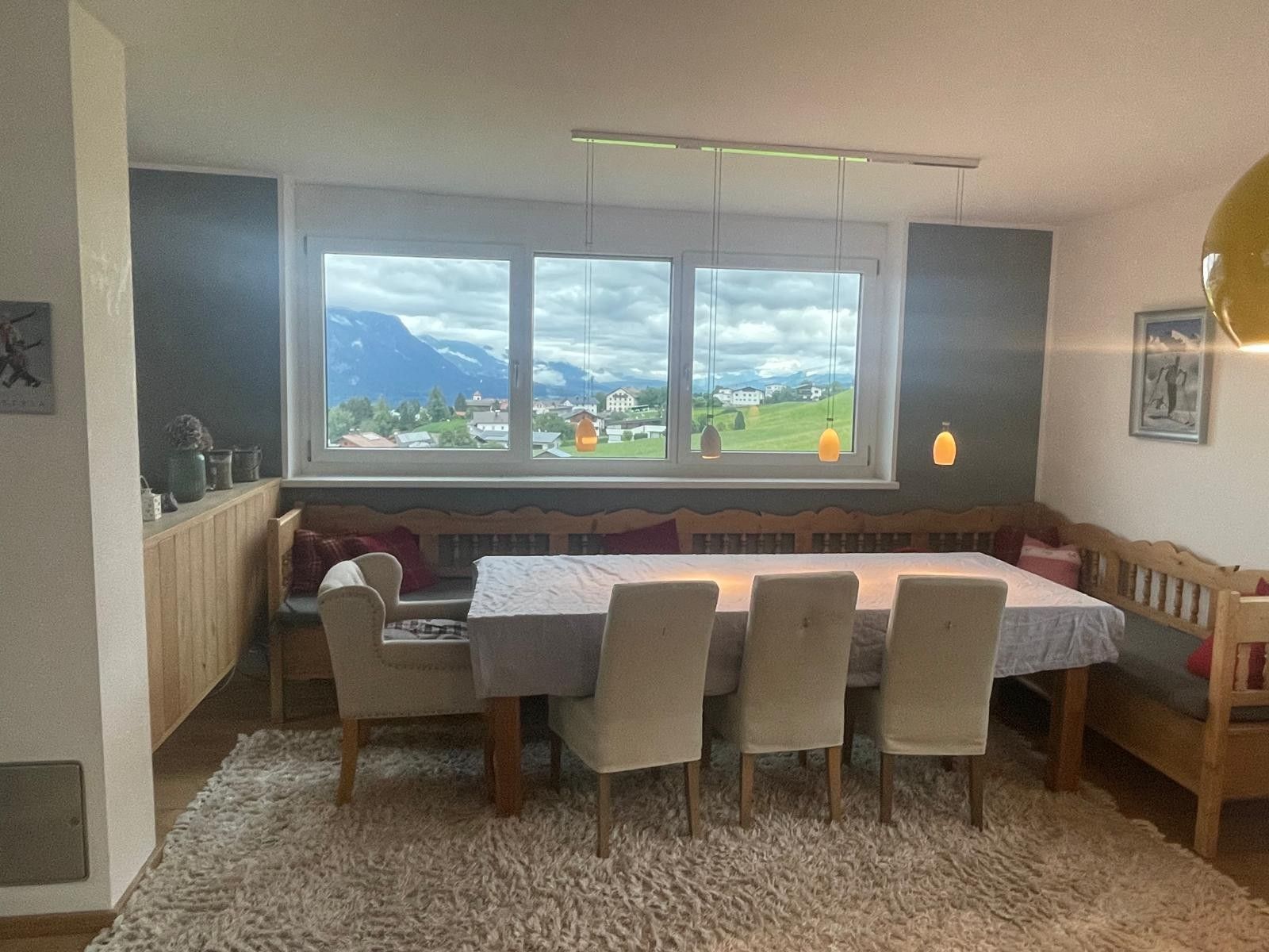 En matplats med ett bord och stolar, en bänk, utsikt över bergen genom stora fönster och en hängande lampa.