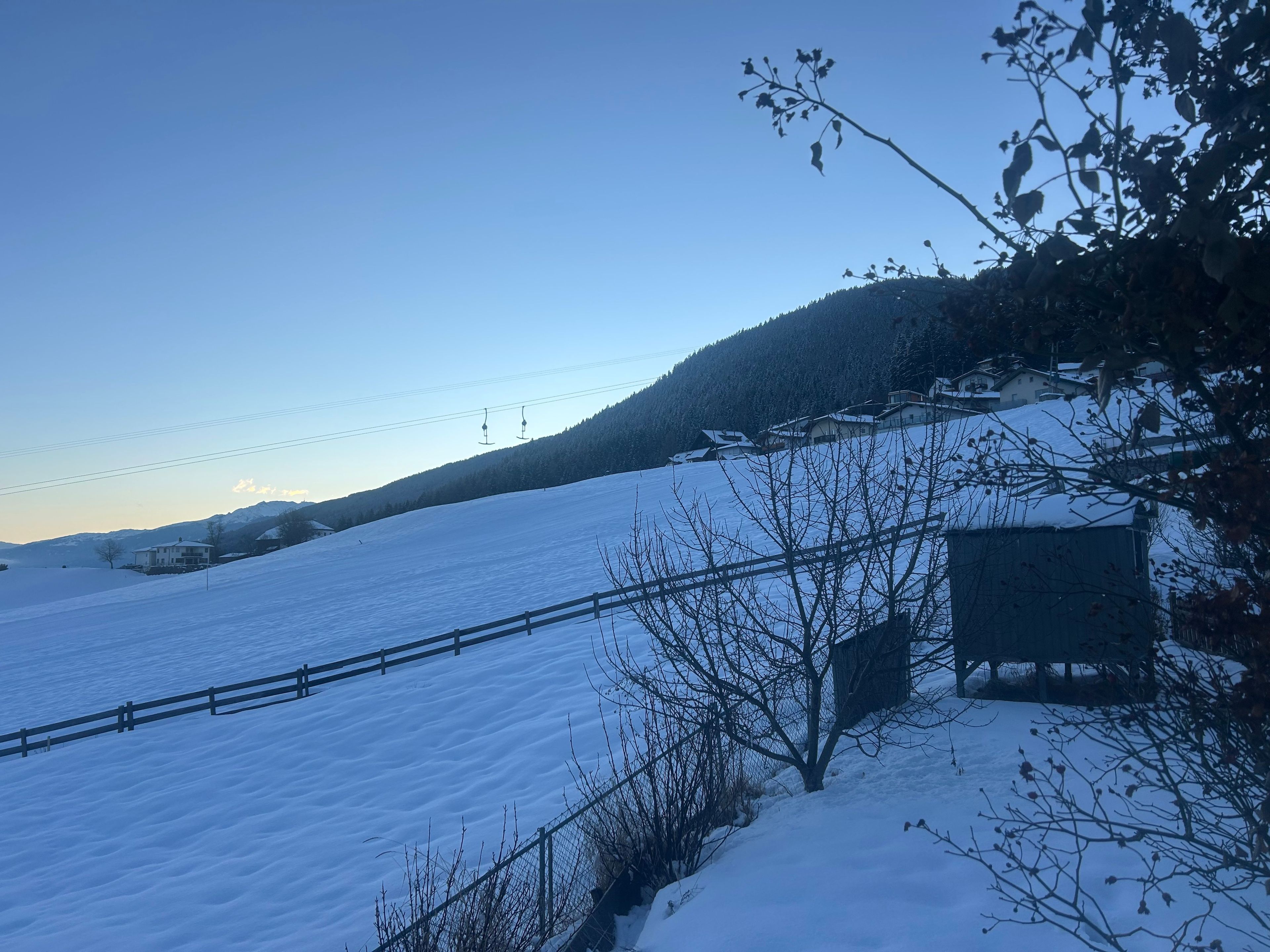 A snow-covered landscape with a wooden fence, a small building, and a hill with houses in the background.
