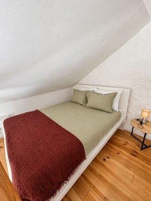 A single bed with a red blanket, a green bedspread, and two pillows in a room with wooden flooring and a white textured wall. - A Window to Porto - Corticeira Lovely House (Porto)
