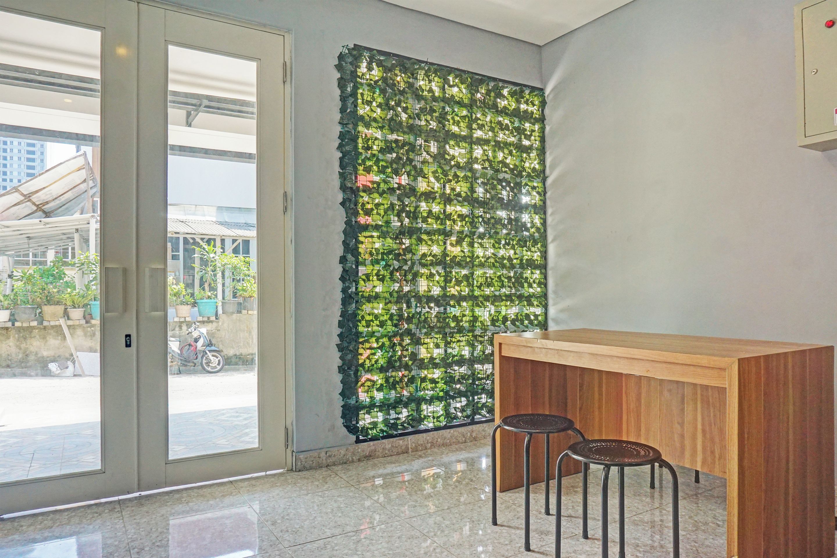 A room with a glass door leading outside, a wooden counter with two stools, and a wall with a green leafy installation.