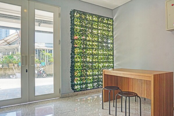 A room with a glass door leading outside, a wooden counter with two stools, and a wall with a green leafy installation.