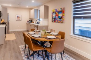 A dining area with a round table set for four, a kitchen in the background, and a large window with blinds. - The Hudson Residence- Luxury 2BR-Parking-Near NYC (Union City)