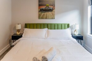 Habitación de hotel con una cama bien hecha, dos mesitas de noche con lámparas, un cabecero tapizado en verde y un cuadro de un coche en la pared.