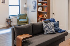 A living room with a grey sofa, a wooden side table, a green armchair, a bookshelf, and a lamp. - The Hudson Residence- Luxury 2BR-Parking-Near NYC (Union City)