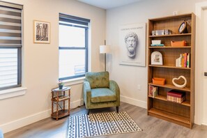 Ein Zimmer mit einem grünen Sessel, einem Holzregal, einem gemusterten Teppich und einem Fenster mit Jalousien.