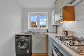 A compact kitchen with a built-in washer, a gas stove, and a window overlooking a garden. - Spacious 3-Bed Home | 4 Mins to Universal Studios | Parking & Fast WiFi (Marston Moretaine)