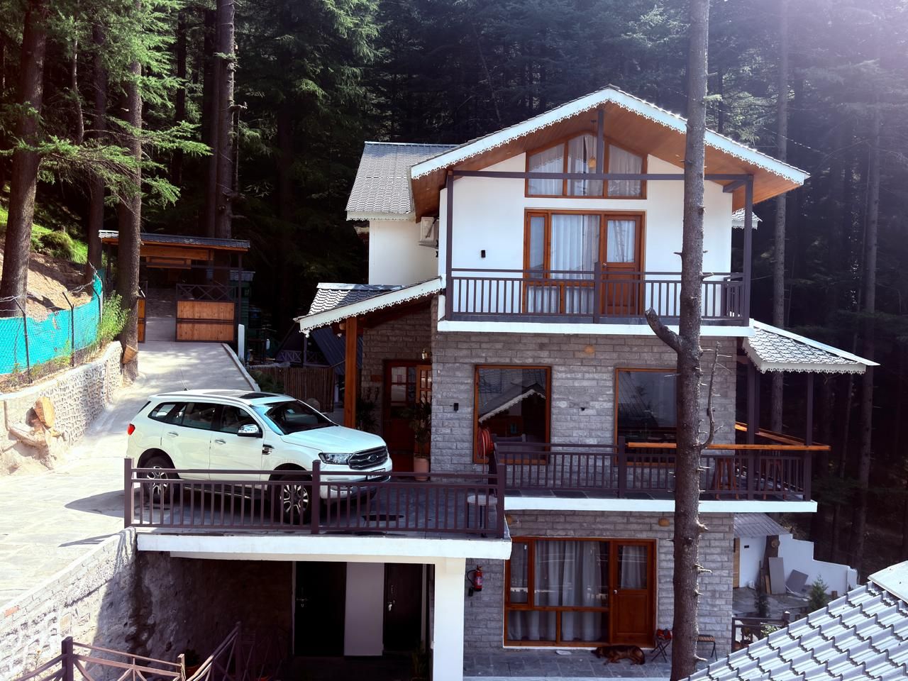 A two-story building with a balcony, a car parked in front, and a forested background.