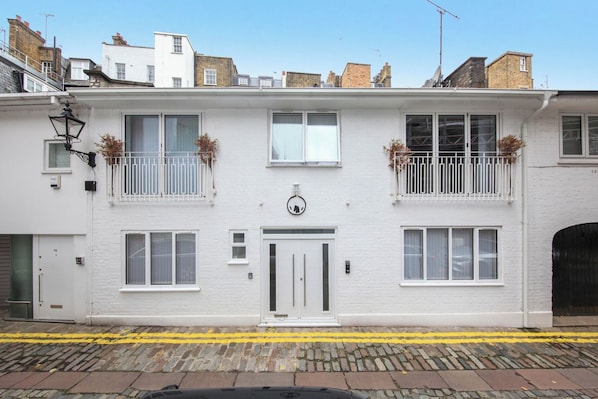 Un edificio blanco de dos pisos con puerta principal, ventanas y un balcón con plantas.