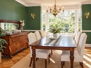 A dining room with a large wooden table, white upholstered chairs, a chandelier, and a view through large windows. - Hopewell Creek Farm - Amish Country Farmhouse, Creek & Fall Color - Sleeps 8 (Lancaster)