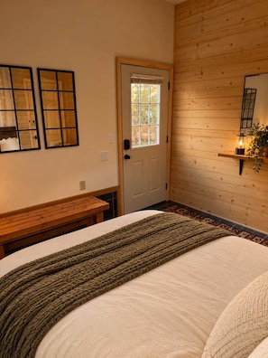 A bedroom with a wooden door, a bench, a bed with a knitted throw, and a wall-mounted shelf with a lamp. - Glamping Cabin 7 | Queen Bed, Trailside, Pets OK (Bessemer Township)