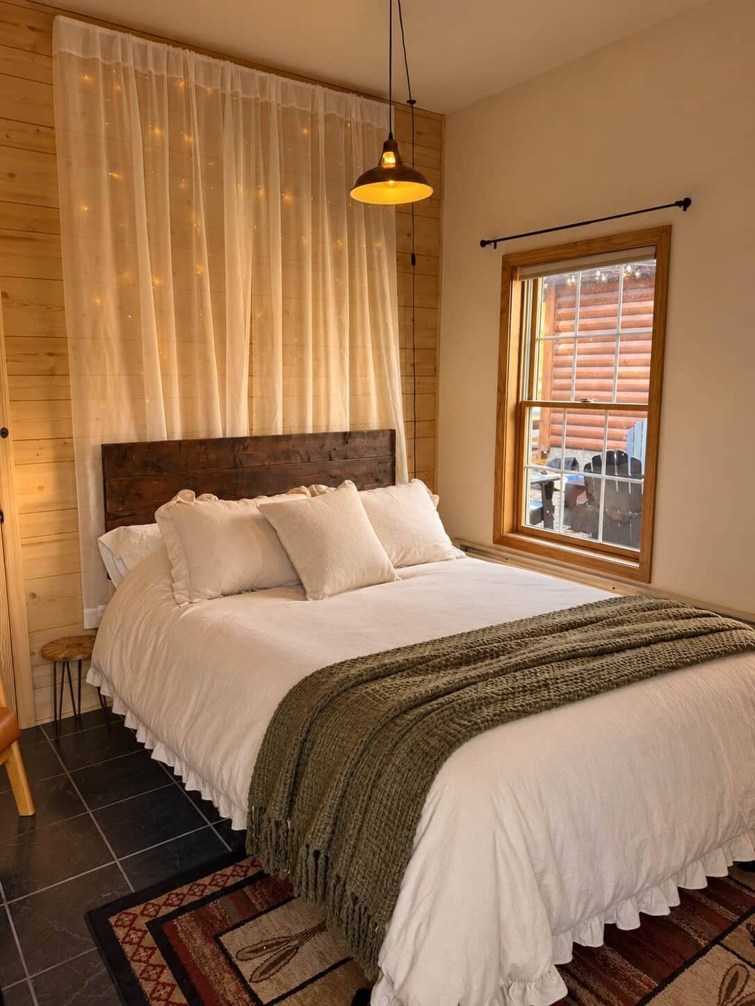 A bedroom with a large bed, a wooden headboard, a window with a view, and a hanging light fixture.