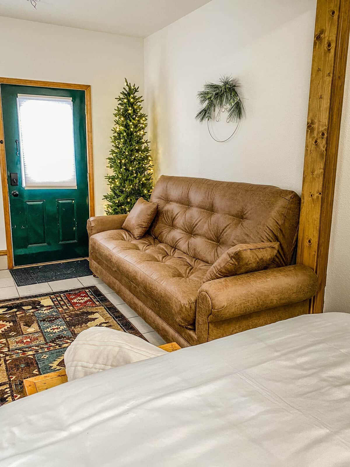 Ein Zimmer mit einem braunen Ledersofa, einem gemusterten Teppich, einem weißen Bett, einer grünen Tür und einem kleinen Weihnachtsbaum.