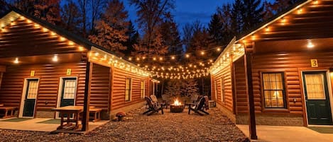A row of log cabins with outdoor seating and a fire pit, surrounded by trees.