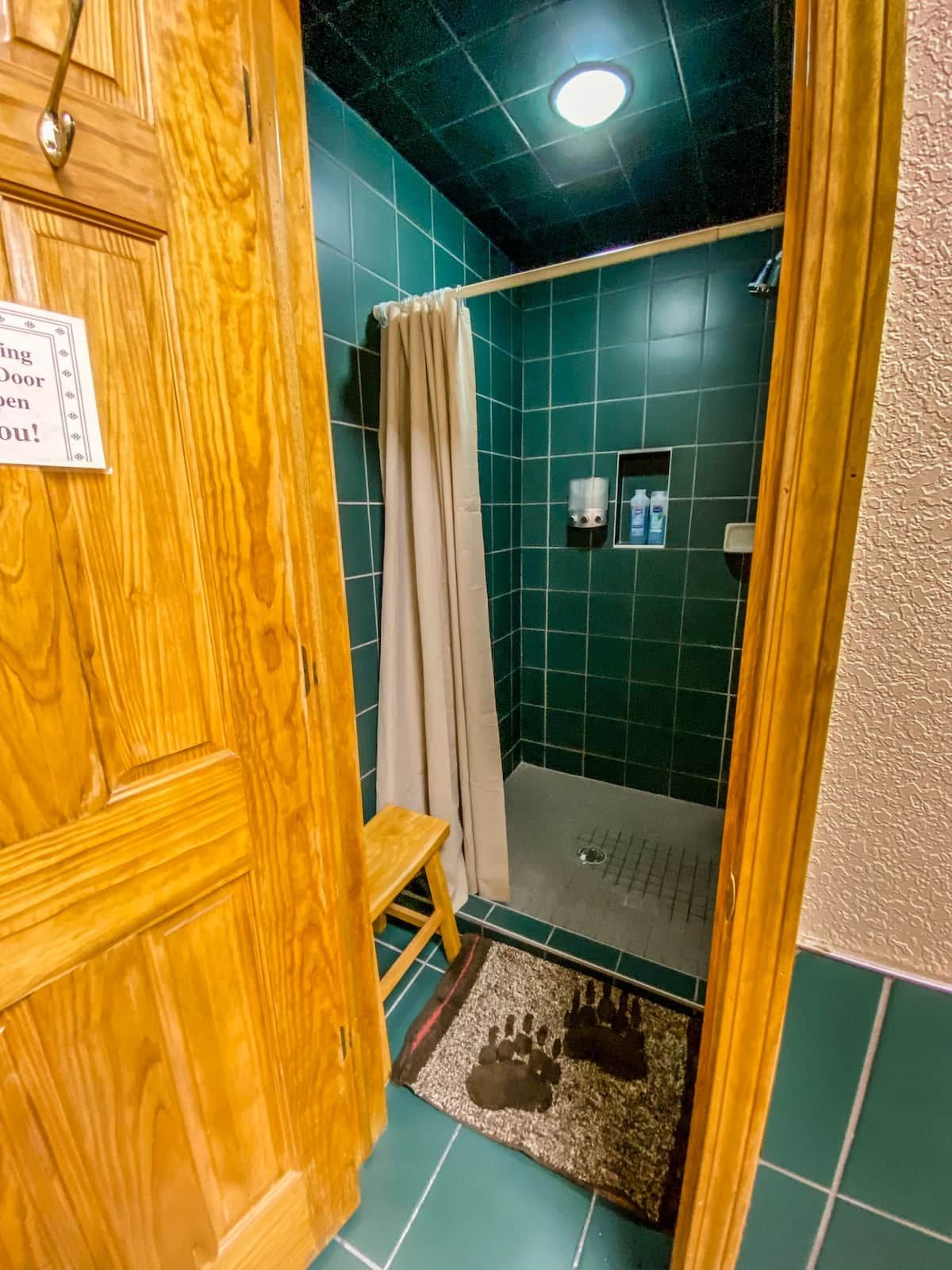A wooden door with a sign, a shower area with green tiles, a beige curtain, and a small shelf with toiletries.