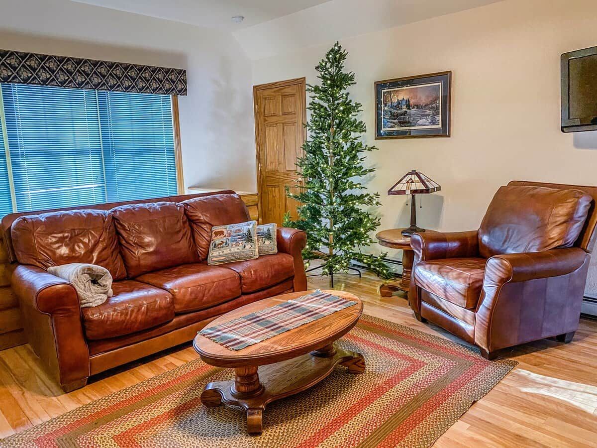 Una sala de estar con un sofá de cuero marrón, un sillón a juego, una pequeña mesa de centro de madera, un árbol de Navidad y un cuadro enmarcado en la pared.