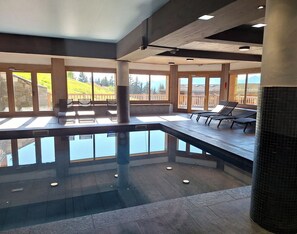 A modern indoor swimming pool with clear glass walls, wooden deck seating, and a view of a mountainous landscape. - Ski-in/ski-out apartment with Mont Blanc view (Bourg-Saint-Maurice)