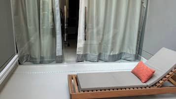 A balcony with a wooden lounge chair and a red pillow, a sliding glass door with sheer curtains, and a tiled floor.