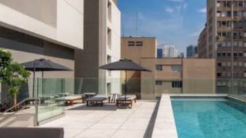 A rooftop pool area with lounge chairs and umbrellas, surrounded by high-rise buildings.