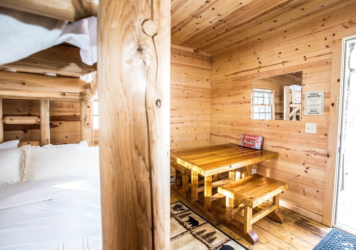 Interior de una cabaña de madera con una cama, una mesa de madera con bancos y una ventana.
