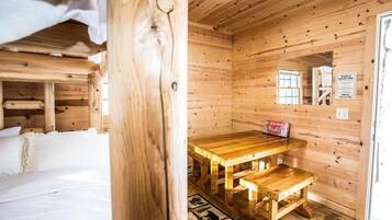 Interior de una cabaña de madera con una cama, una mesa de madera con bancos y una ventana.