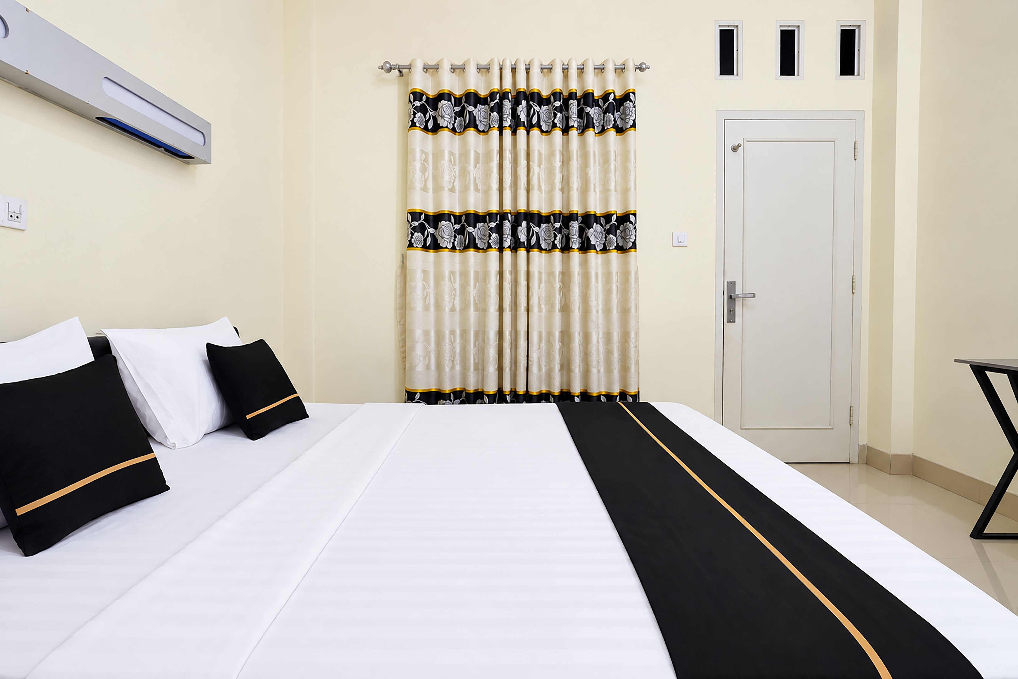 A hotel room with a bed, pillows, a curtain, a door, and an air conditioning unit.