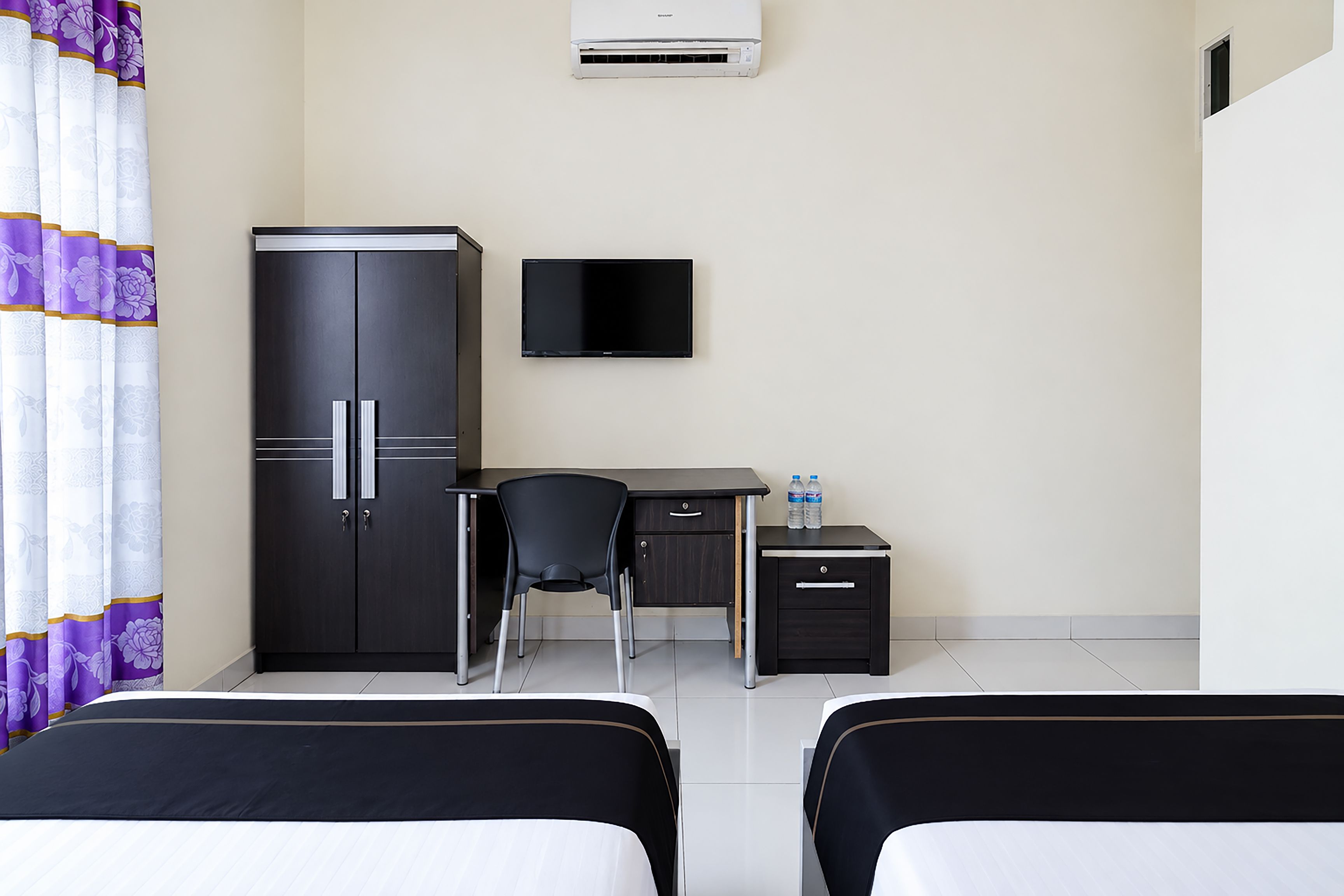 A hotel room with two beds, a desk, a chair, a wardrobe, a television, and a water bottle.