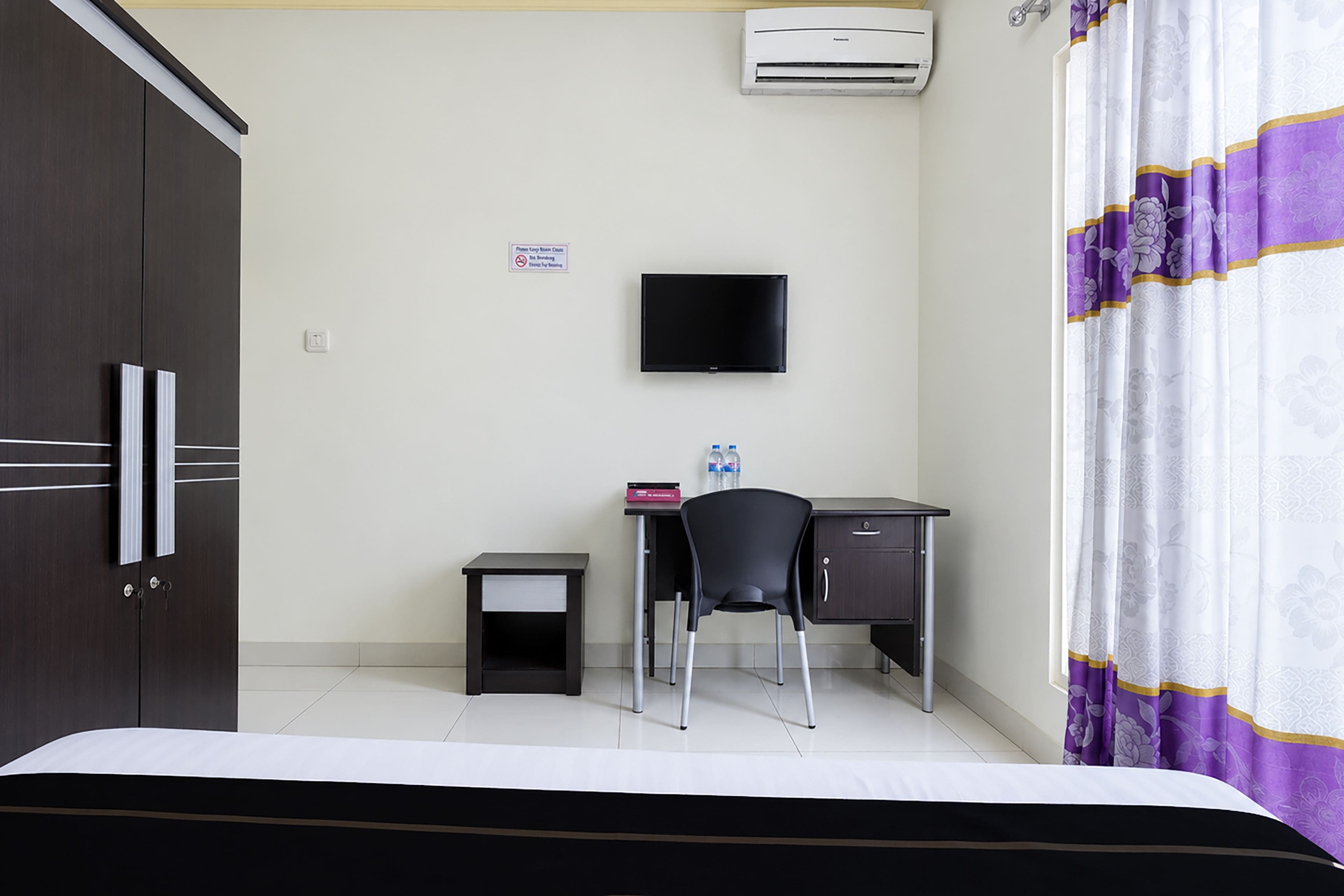 Een hotelkamer met een bed, een bureau met stoel, een televisie, een airconditioner en een gordijn.