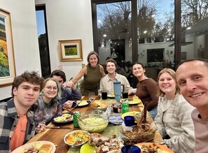 Un grupo de personas sentadas alrededor de una mesa con diversos platos, en una habitación con ventanas amplias y cuadros en las paredes.