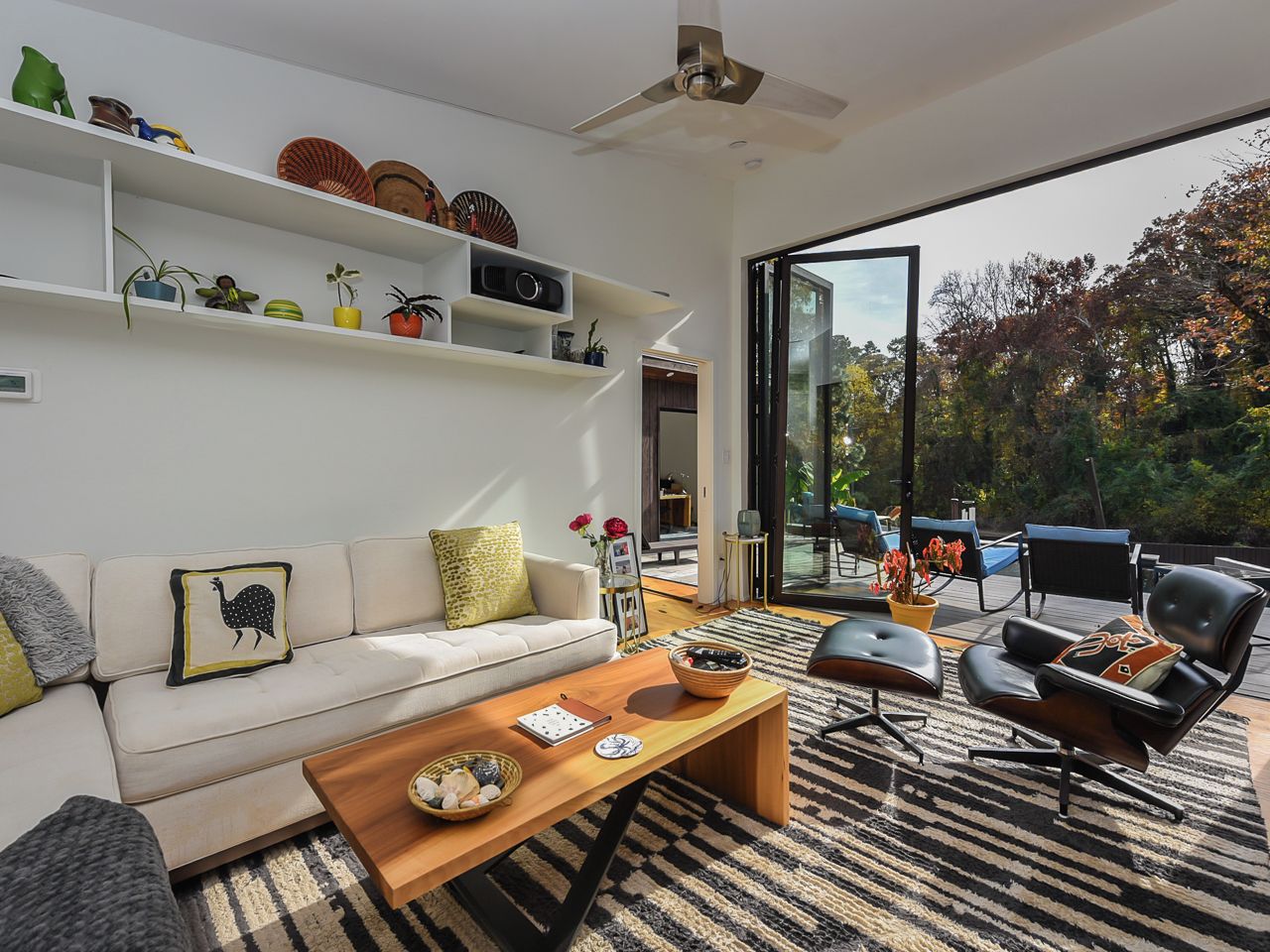 A modern living room with a sofa, coffee table, and chairs, a bookshelf with decorative items, and a large glass door leading to an outdoor area.