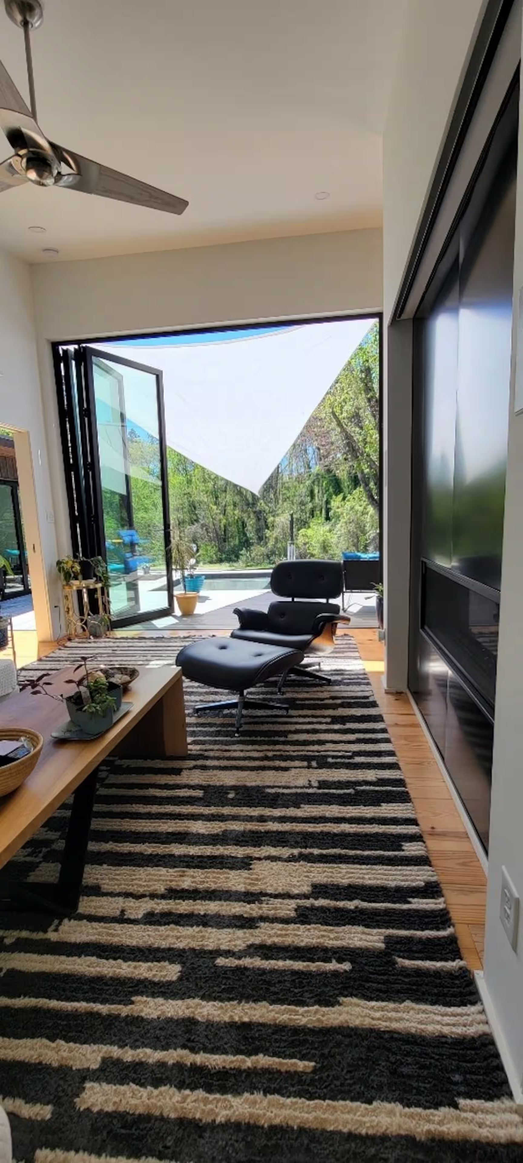 A modern living room with a large flat-screen TV, a ceiling fan, and a view of a pool and greenery.