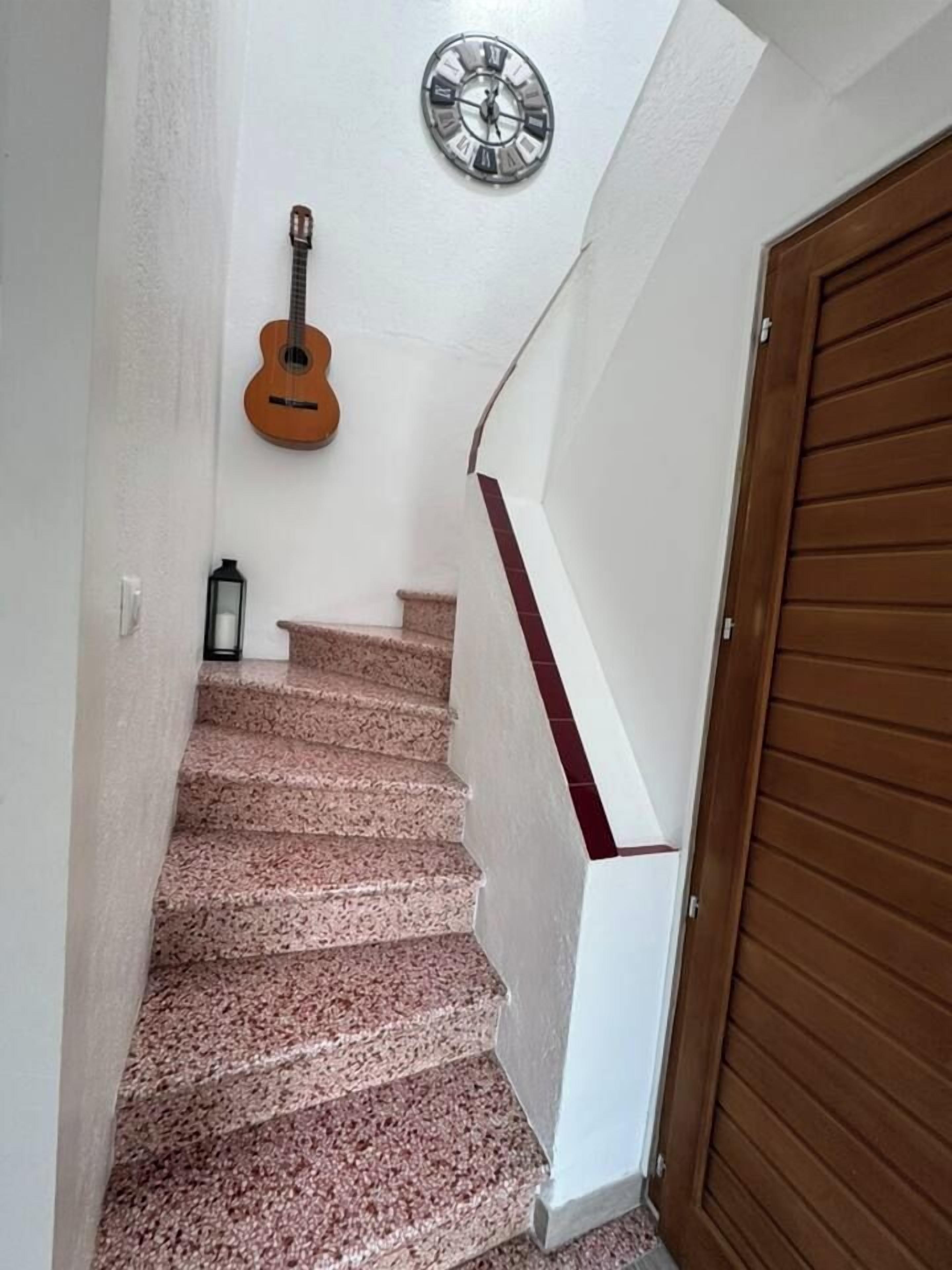 Eine Treppe mit hölzernem Geländer, einer an der Wand befestigten Uhr und einer an die Wand gelehnten Gitarre.