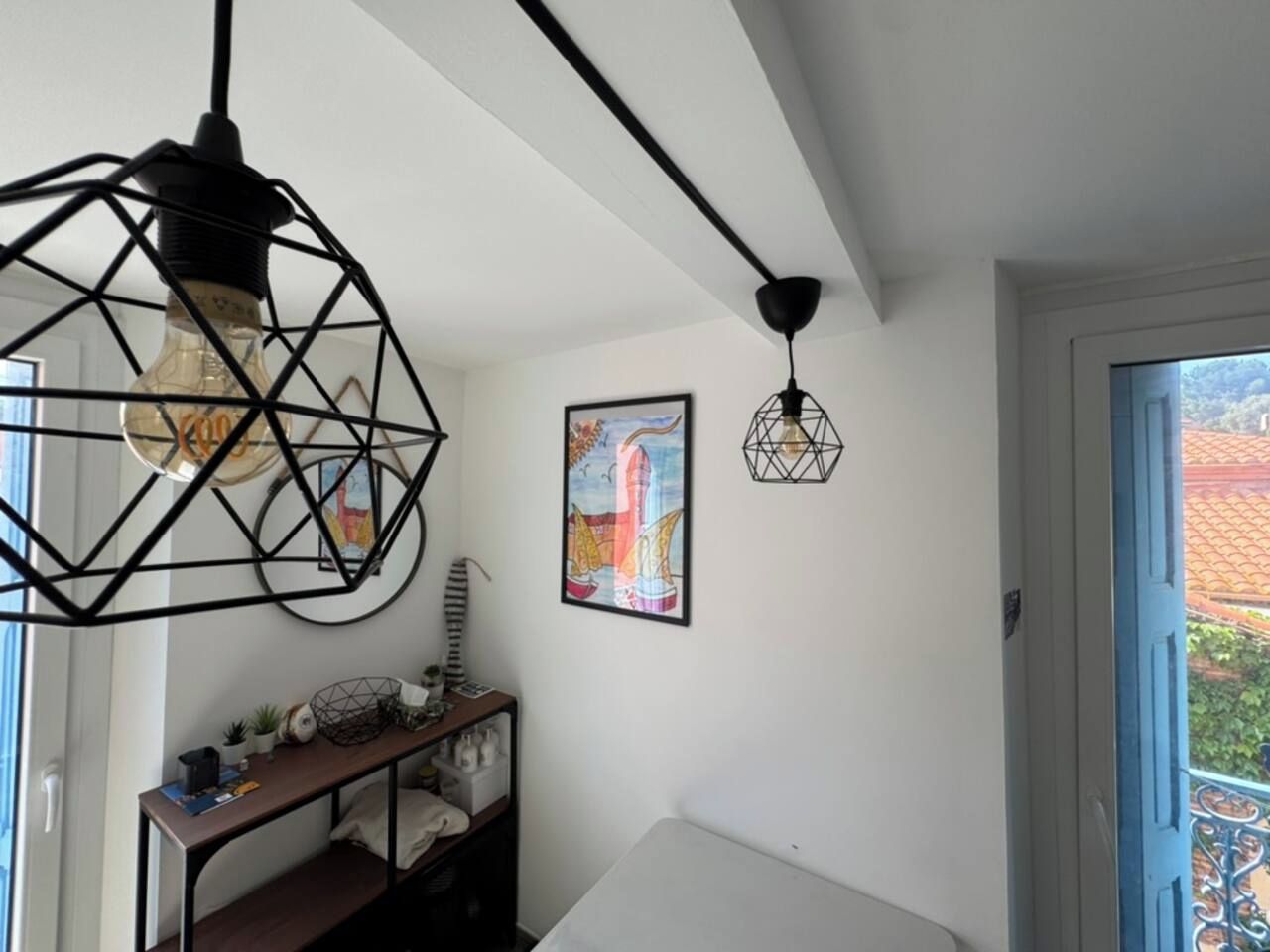 A room with a white wall, a framed colorful painting, a small table with decorative items, and a door leading to an outdoor area.