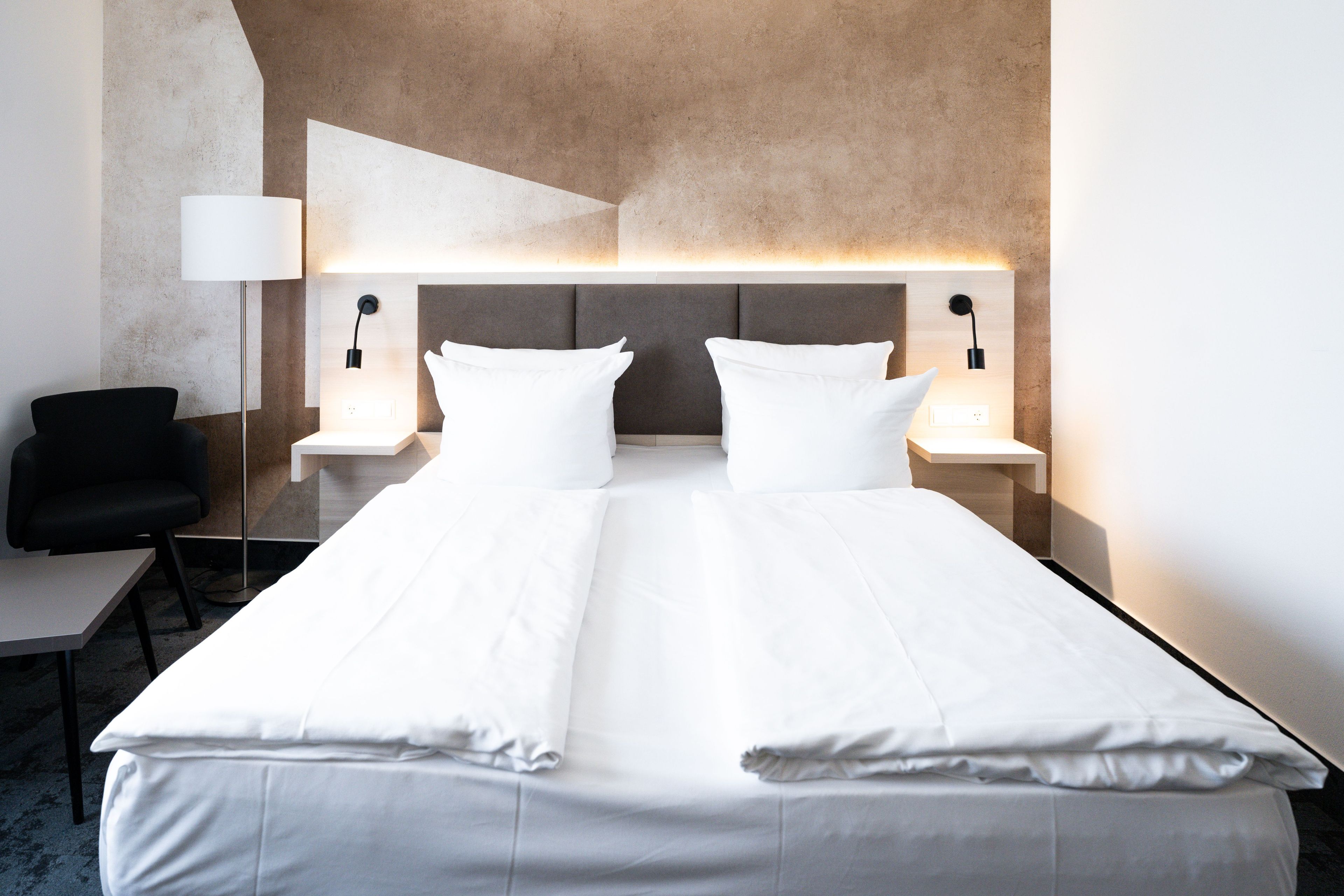 A modern hotel room with a large bed, two bedside tables with lamps, a chair, and a desk.