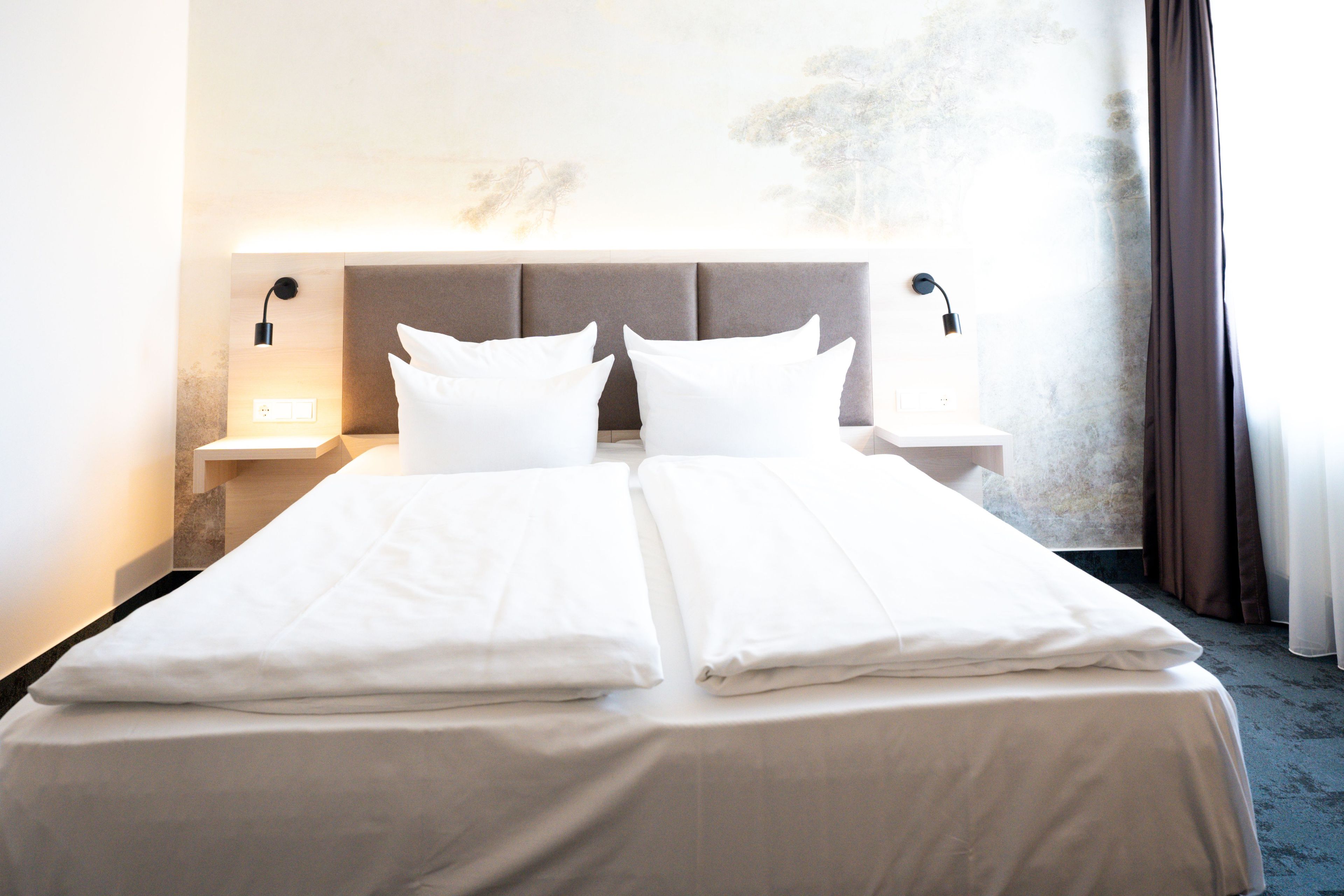 A double bed with two pillows, a headboard, bedside tables with lamps, and a view of the outdoors through a window.