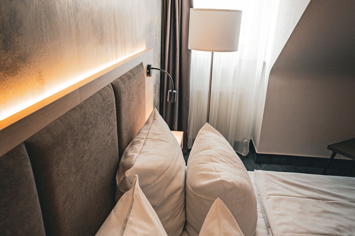 Una habitación de hotel con una cama, almohadas, una lámpara y un cabecero.