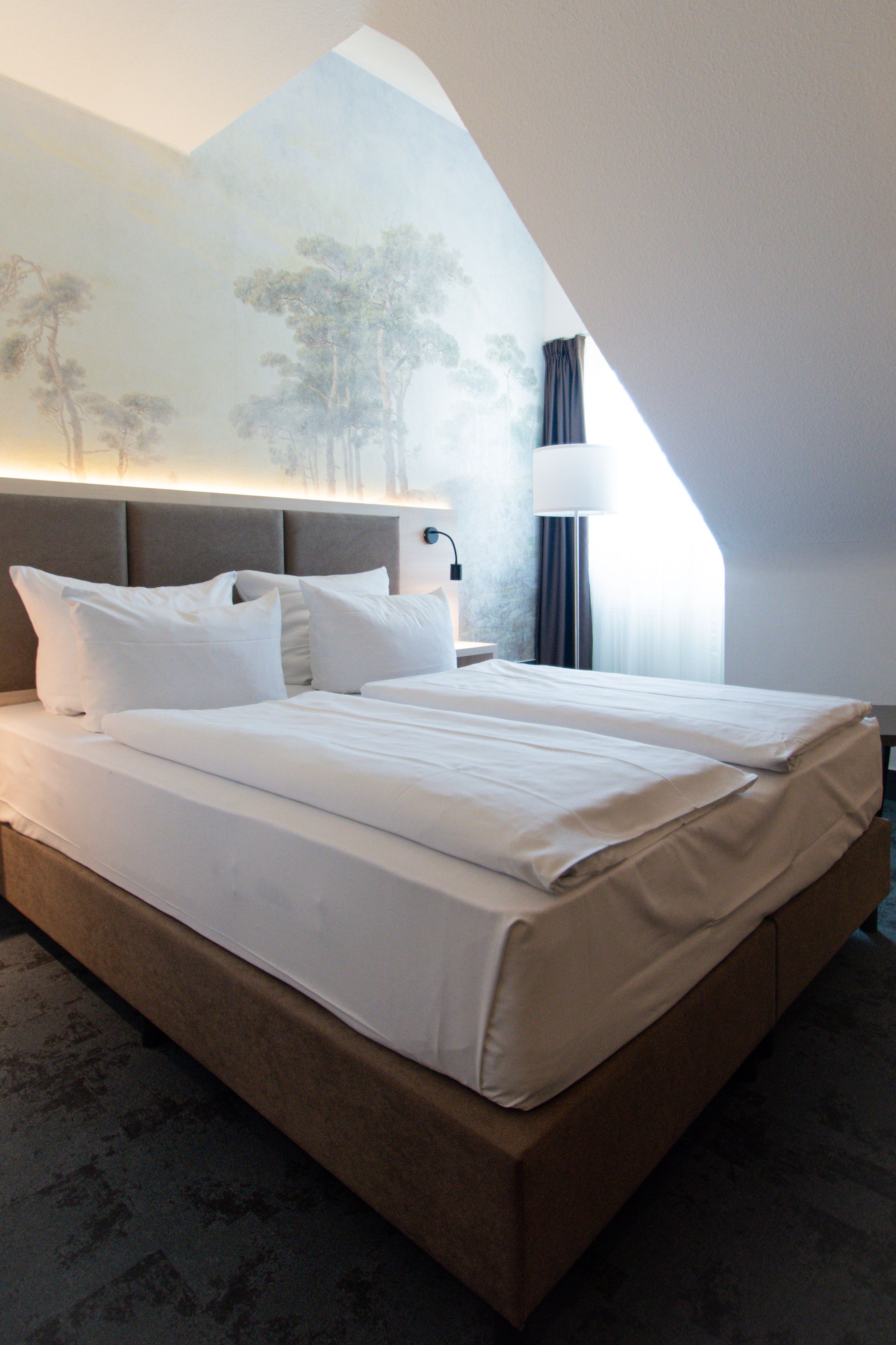 A hotel room with a large bed, a nightstand, a lamp, and a wall mural.
