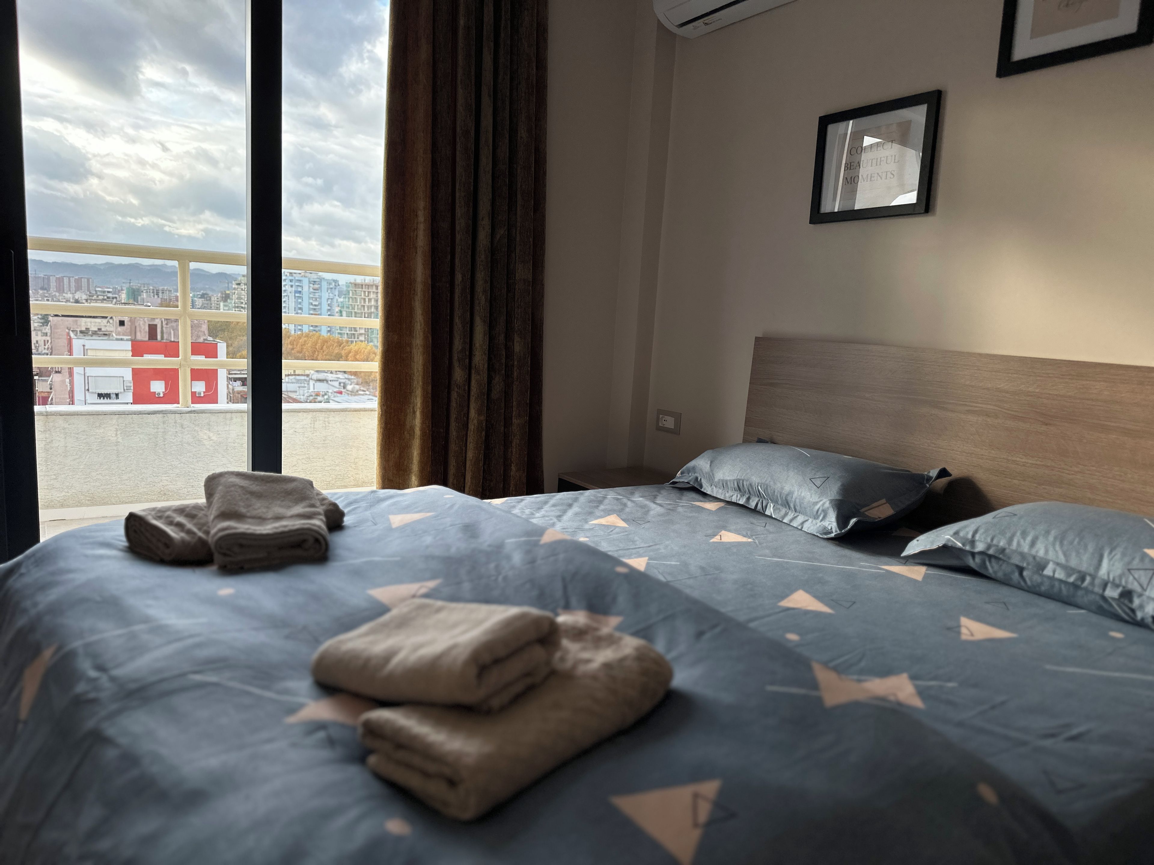 A hotel room with a bed, a window with curtains, a framed picture on the wall, and a view of the cityscape.