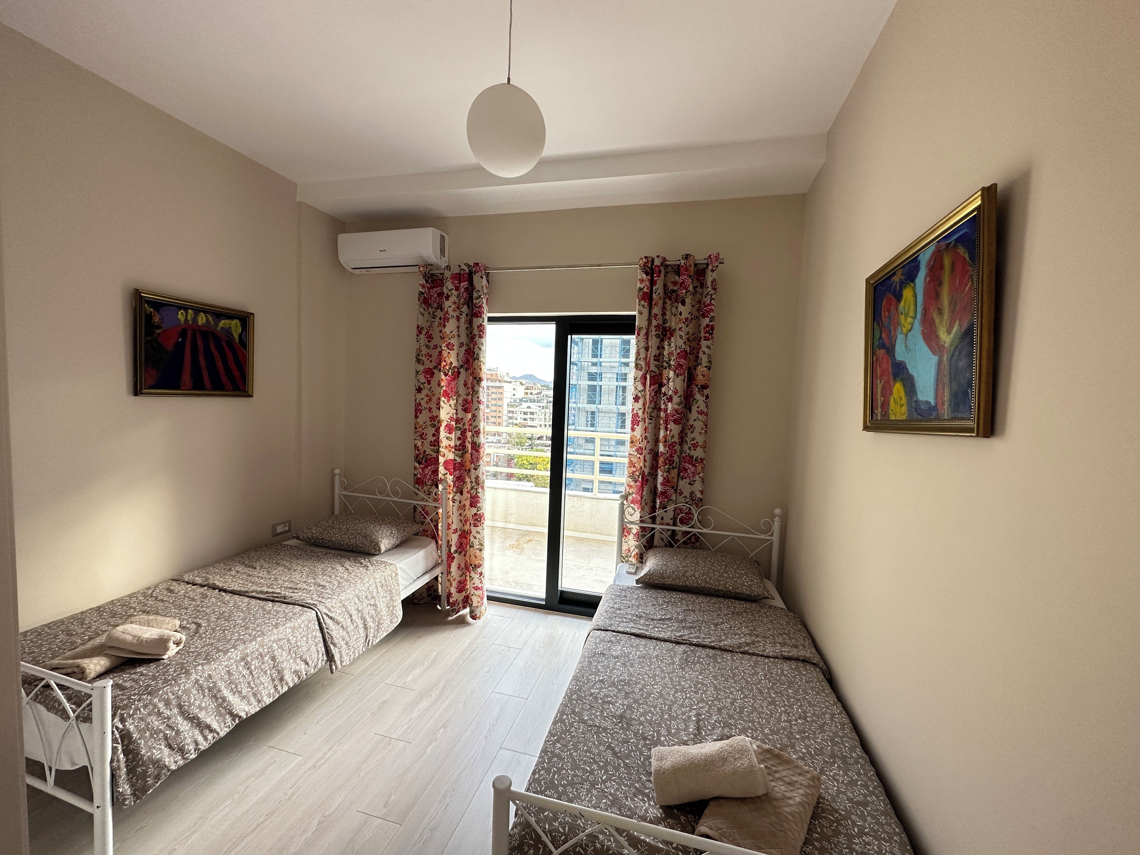 A room with two beds, a ceiling fan, a window with curtains, and a painting on the wall.