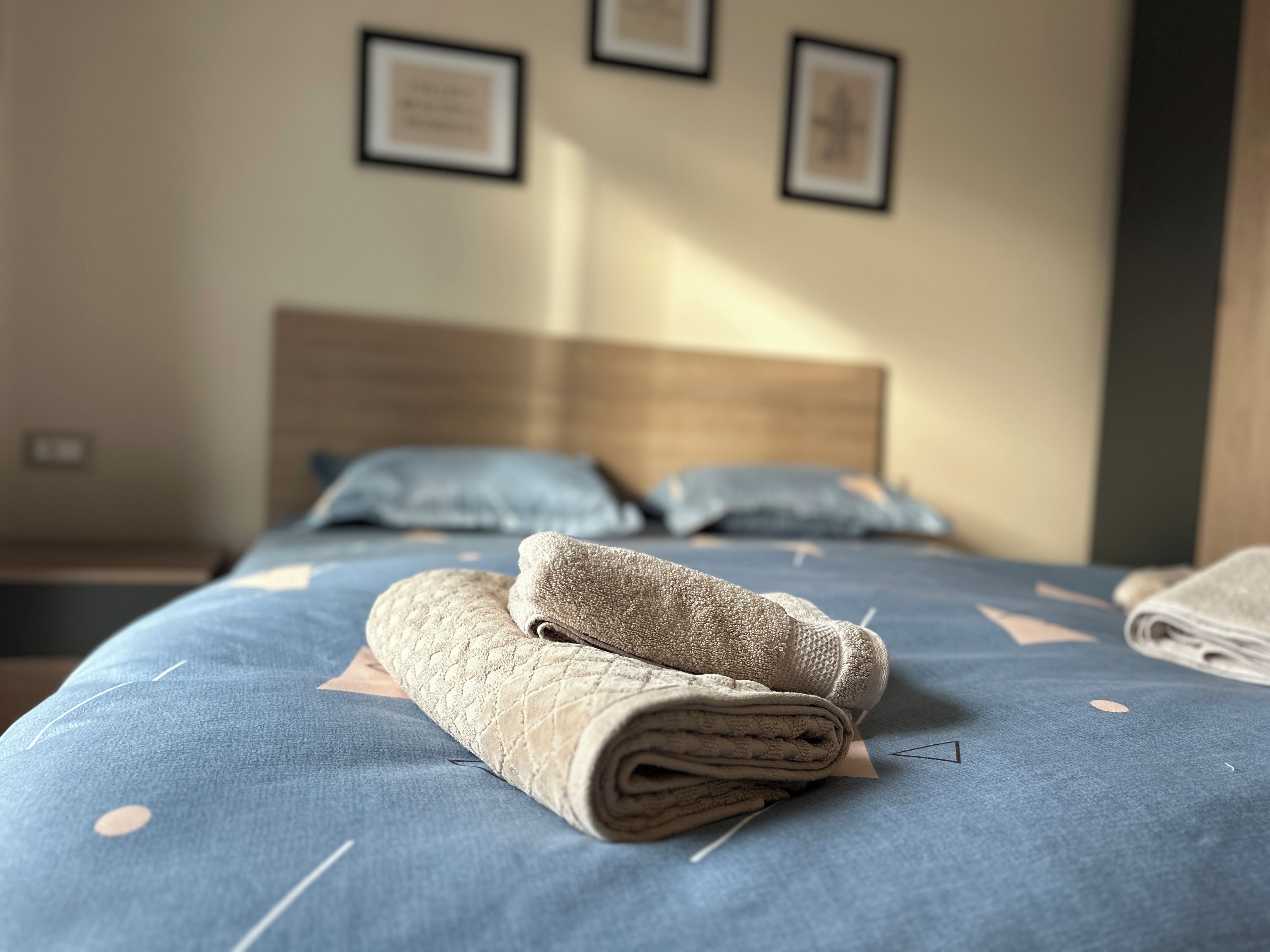 A neatly made bed with a blue bedspread and two pillows, a folded blanket on the bed, and a wooden headboard.