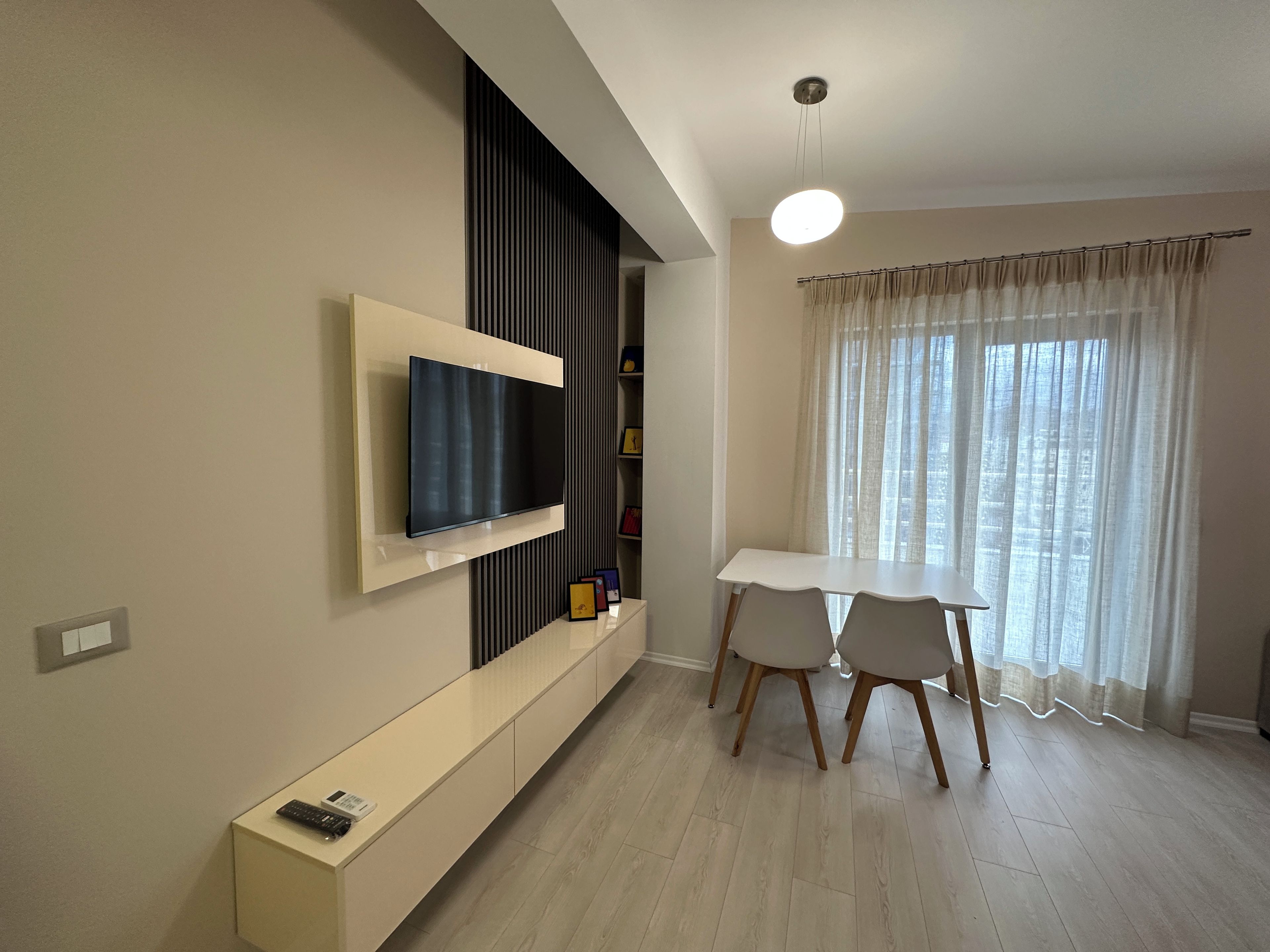 A modern living room with a flat-screen TV mounted on the wall, a dining table with chairs, and a large window with sheer curtains.