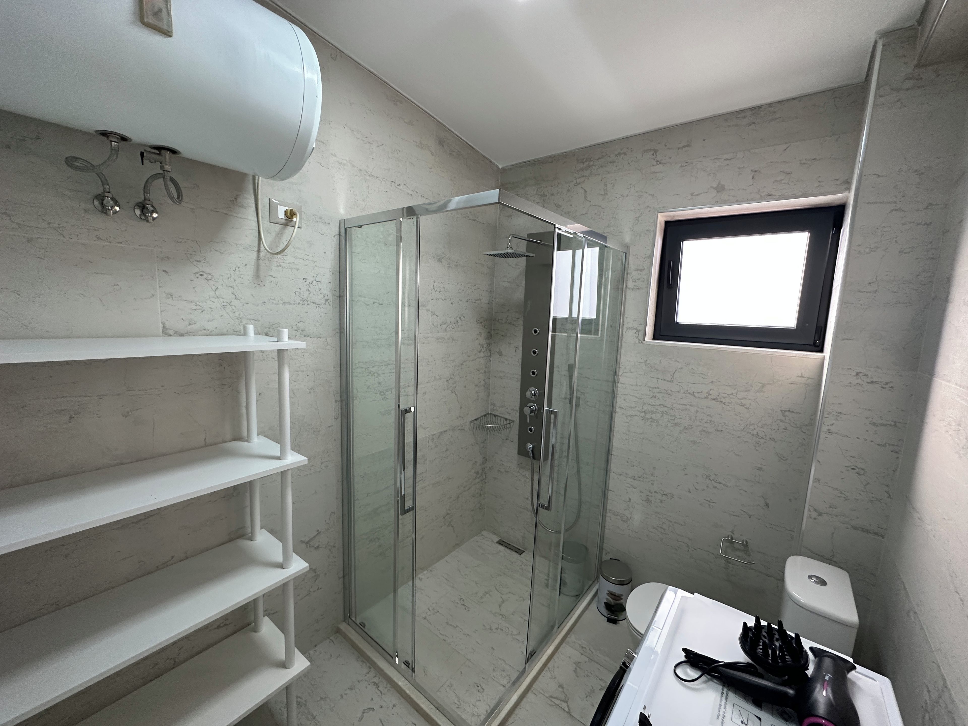 Un baño moderno con ducha de vidrio, inodoro y lavamanos, además de un secador de pelo y otros artículos de aseo.