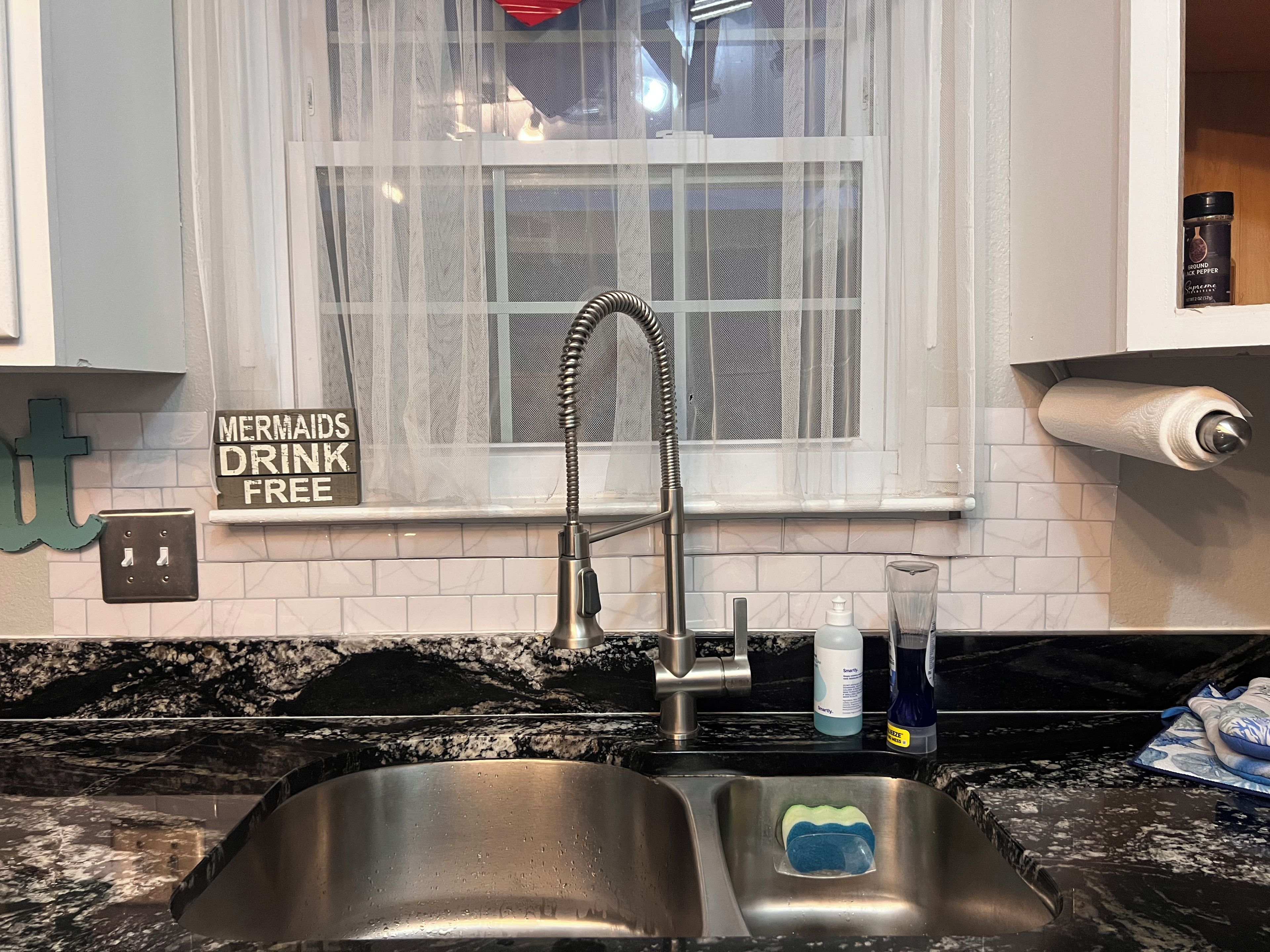 A kitchen with a stainless steel sink, a window with a white curtain, and a sign that says 'Mermaids Drink Free.'