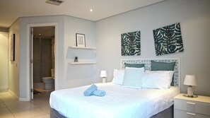 A modern bedroom with a large bed, two wall-mounted artworks, a bedside table with a lamp, and a small shelf with a plant. - 2-bedroom apartment in charming Durban with AC for relaxing stays (Durban)