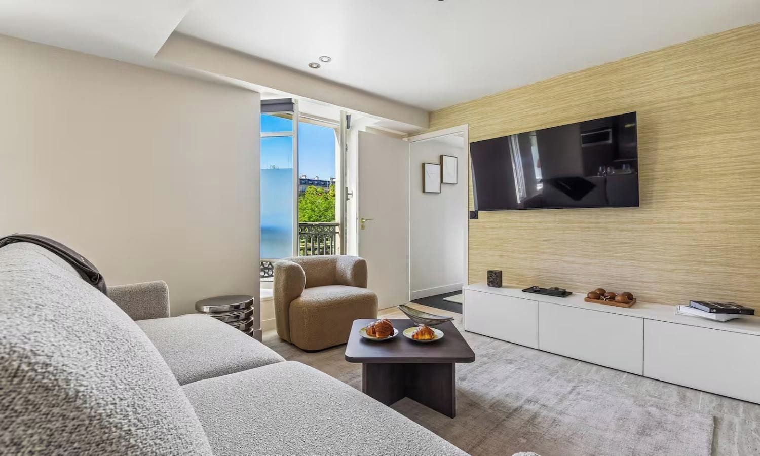 Un salon moderne avec un canapé, un fauteuil, une petite table sur laquelle se trouve une assiette de nourriture, un téléviseur à écran plat fixé au mur et une porte coulissante donnant sur un balcon avec vue.