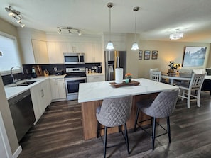 A modern kitchen with stainless steel appliances, a marble island, and a dining area with a view of the mountains. - Family & Pet Friendly 3BR, Large Fenced Yard- Close to South Health Campus (Calgary)