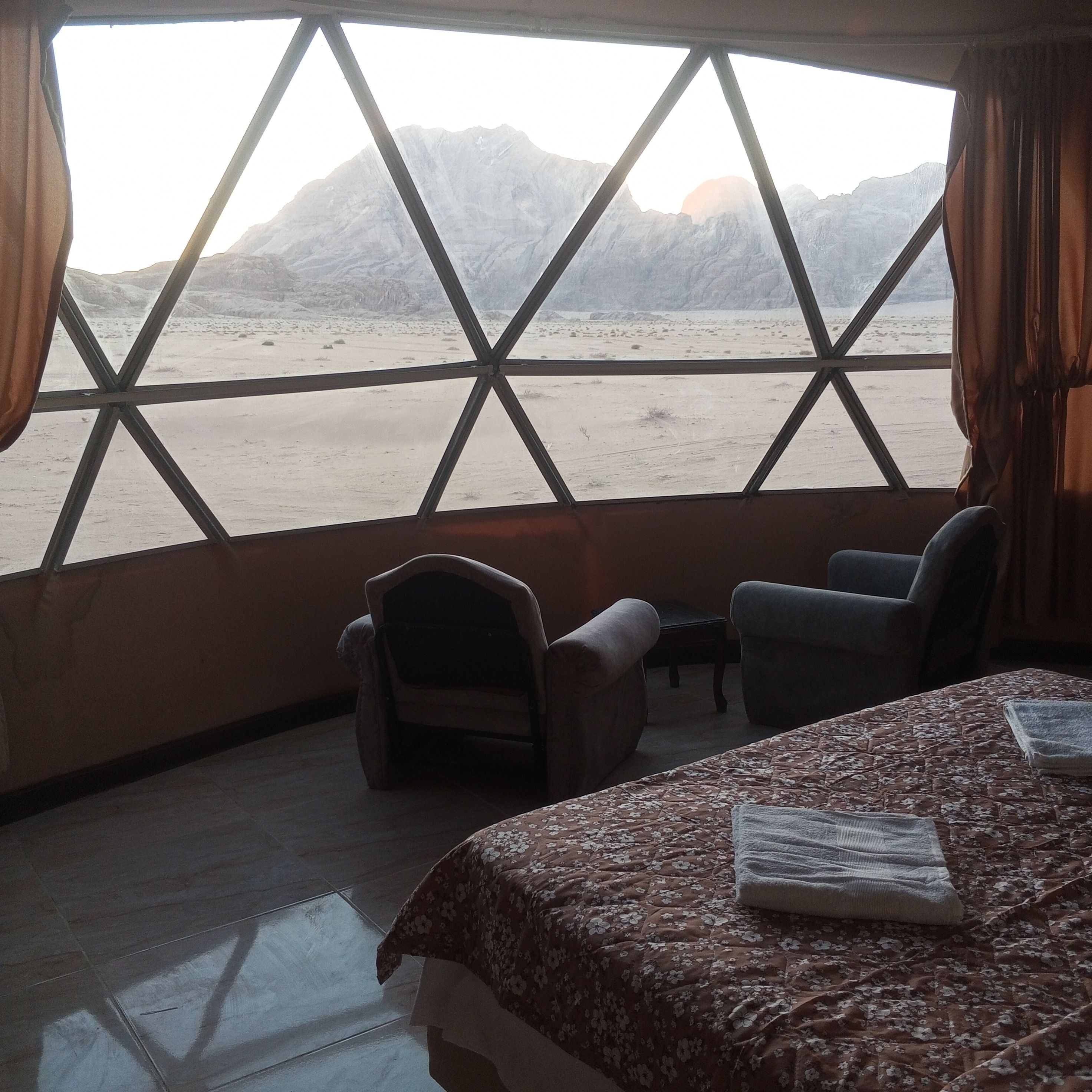 Ein Zimmer mit einem Bett, zwei Stühlen und einem großen Fenster mit Blick auf eine Berglandschaft.