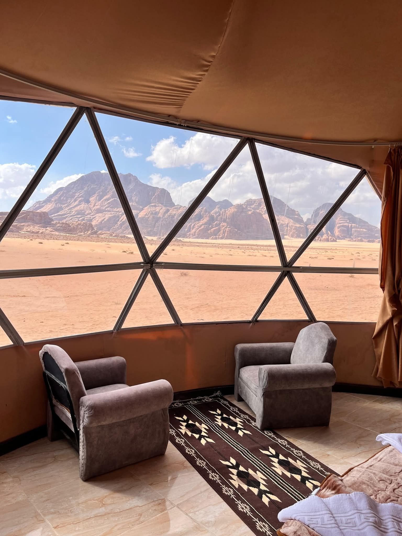 A tent with a view of a desert landscape and mountains. Inside, there are two chairs and a patterned rug.